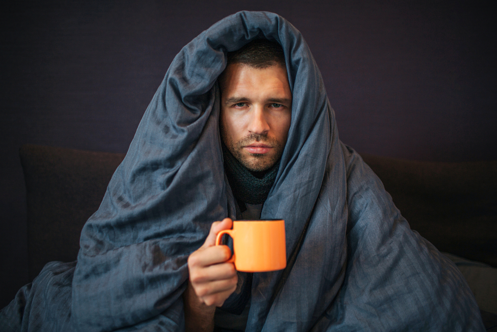 Man with sad look on face under doona holding coffee mug