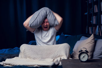 Man sitting in bed with pillow over his head at 1:37am