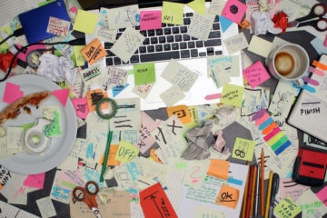 Desk with laptop covered in post-it notes, scissors, coffee cups and plate with old pizza